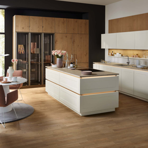 Moderne Landhausküche mit Kochinsel, Kehlleistenbeleichtung und grauer Steinarbeitsplatte, sowie Nischenrückwand in Holzoptik und Holzvitrine – Küchen in Marl